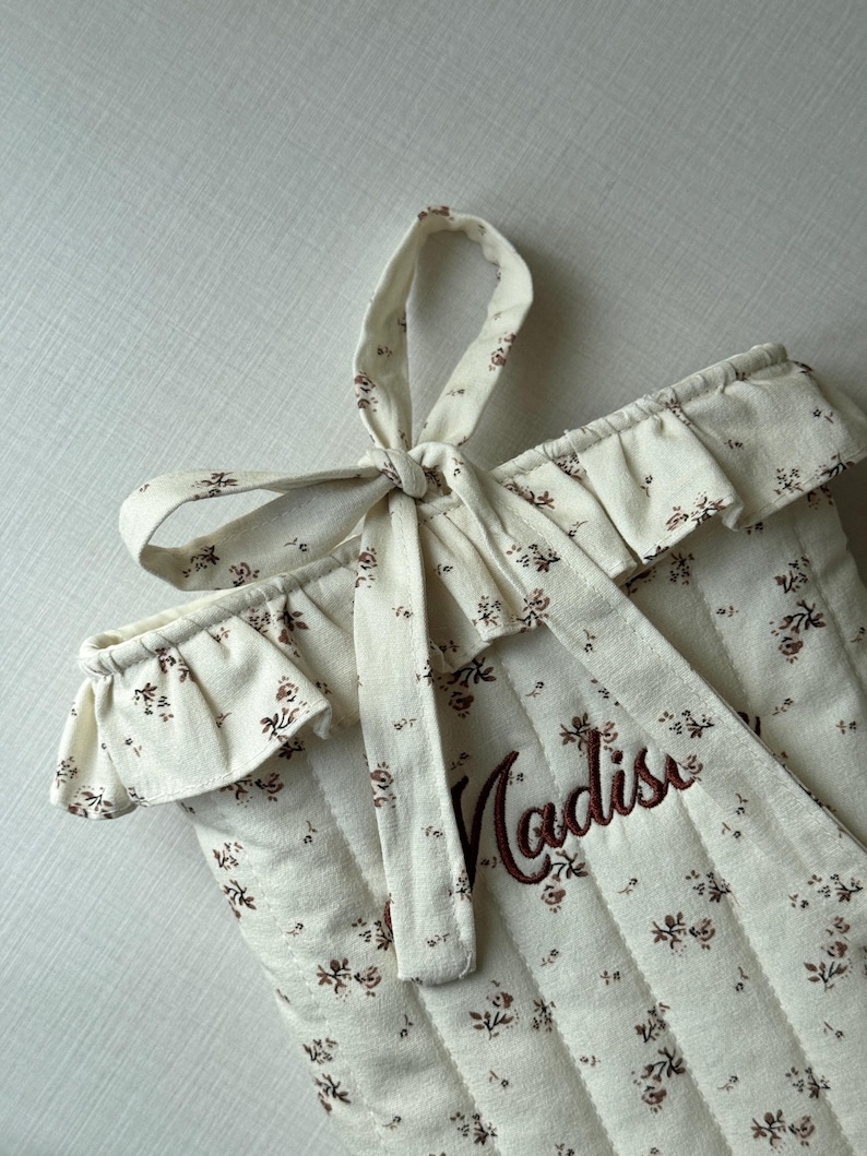 Puede incluir: Tela acolchada de color crema con un estampado floral y el nombre "Madison" bordado en marr&oacute;n. El borde superior tiene un ribete con volantes y un lazo de tela a juego atado en la parte superior. El dise&ntilde;o general es suave y delicado.