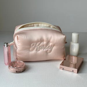 May include: A light pink satin cosmetic bag with the name "Karley" embroidered in a cursive font. The bag is surrounded by makeup items, including a lip gloss, compact, and a bottle of skincare product.