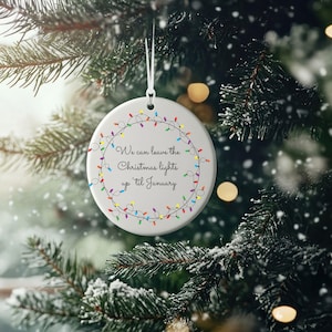 May include: A circular white Christmas tree decoration featuring a garland of multi-coloured lights and the text "We can leave the Christmas lights up 'til January". The ornament hangs from a white string.