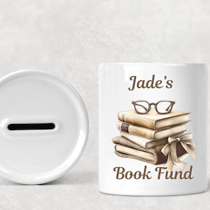 Peut inclure: Tirelire en céramique blanche avec une fente sur le dessus. Le devant de la tirelire est orné d'une illustration brune d'une pile de livres surmontée de lunettes et du texte "Jade's Book Fund".