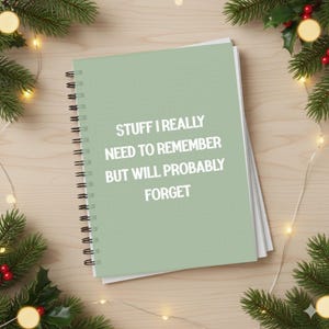 Puede incluir: Un cuaderno de espiral verde claro con el texto "STUFF I REALLY NEED TO REMEMBER BUT WILL PROBABLY FORGET" en blanco. El cuaderno está rodeado de decoraciones navideñas, incluyendo luces y ramas de pino.
