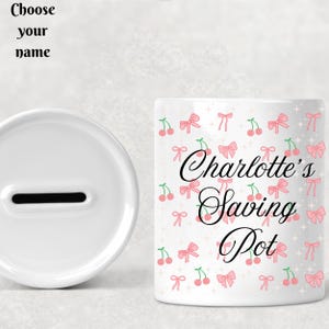 Könnte beinhalten: Weiße Keramik-Sparbüchse mit einem Schlitz oben. Die Vorderseite der Sparbüchse hat ein rosa-weißes Kirsch- und Schleifenmuster mit dem Text "Charlotte's Saving Pot".