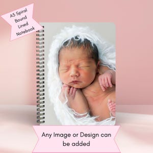 Peut inclure: Un carnet à spirales avec une couverture blanche représentant une photo d'un bébé endormi enveloppé dans une couverture blanche. Le carnet est de format A5 et comporte des pages lignées. Le texte "Any Image or Design can be added" est imprimé sous la photo.