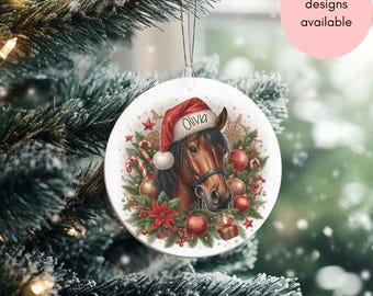 May include: A white, round Christmas ornament featuring a brown horse wearing a Santa hat with the name "Olivia." The ornament is decorated with Christmas elements like candy canes, red ornaments, and greenery. A pink circle with the text "Lots of designs available" is in the background.