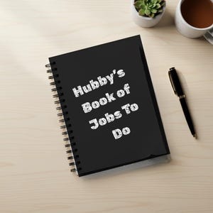Peut inclure: Un cahier noir à spirales avec le texte "Hubby's Book of Jobs To Do" en blanc, écrit à la main. Le cahier est posé sur une surface claire imitation bois, avec un stylo, une petite plante succulente et une tasse de café à proximité.