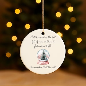 May include: A circular Christmas ornament with a gold hanging cord. The ornament displays a snow globe illustration with a Christmas tree and the text "I still remember the first fall of snow and how it glistened as it fell. I remember it all too well."