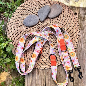 Puede incluir: Dos correas para perros coloridas con un patrón floral y caras sonrientes. Las correas están hechas de tela blanca con flores rosas, naranjas y amarillas. Las correas tienen clips metálicos negros y están sobre un fondo marrón tejido.