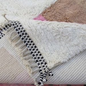 May include: Close-up of a white, fluffy rug with a black and white fringe. The rug has a geometric pattern with pink and brown accents.
