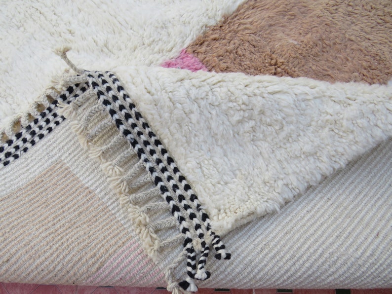 May include: Close-up of a white, fluffy rug with a black and white fringe. The rug has a geometric pattern with pink and brown accents.