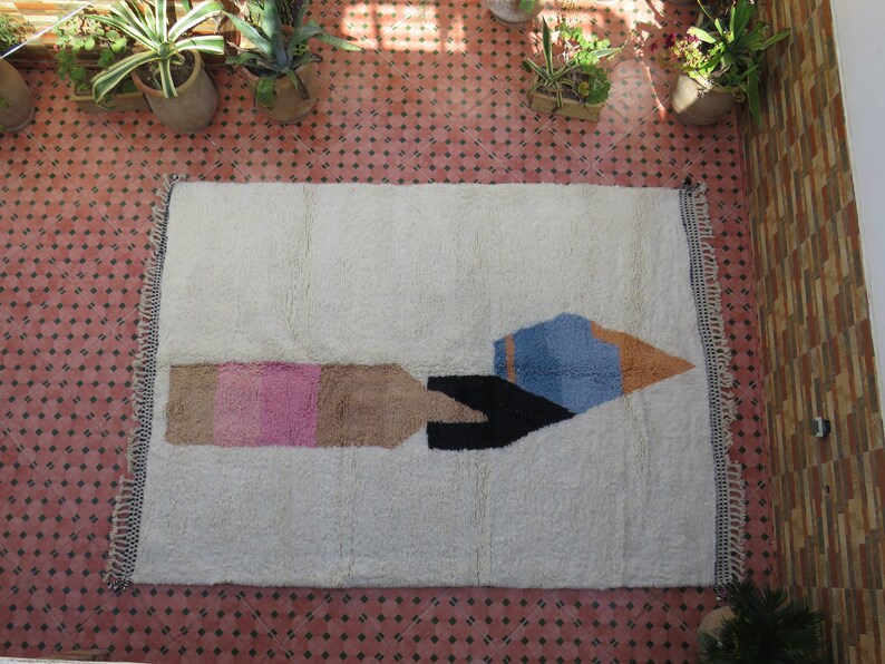 May include: A white rug with a geometric design in pink, brown, black, blue, and orange. The design resembles a stylized rocket ship.