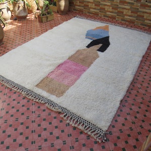 May include: White rug with a geometric design in pink, beige, blue, and black. The rug has fringe around the edges.