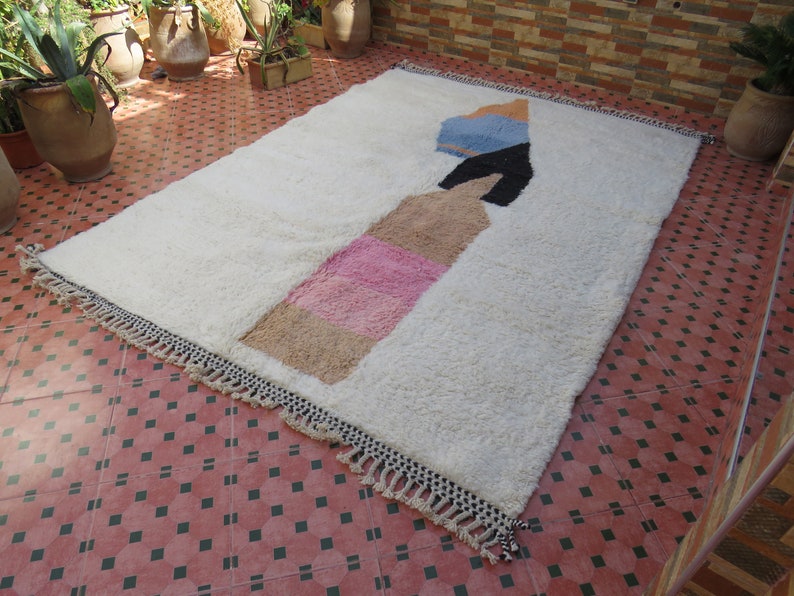 May include: White rug with a geometric design in pink, beige, blue, and black. The rug has fringe around the edges.