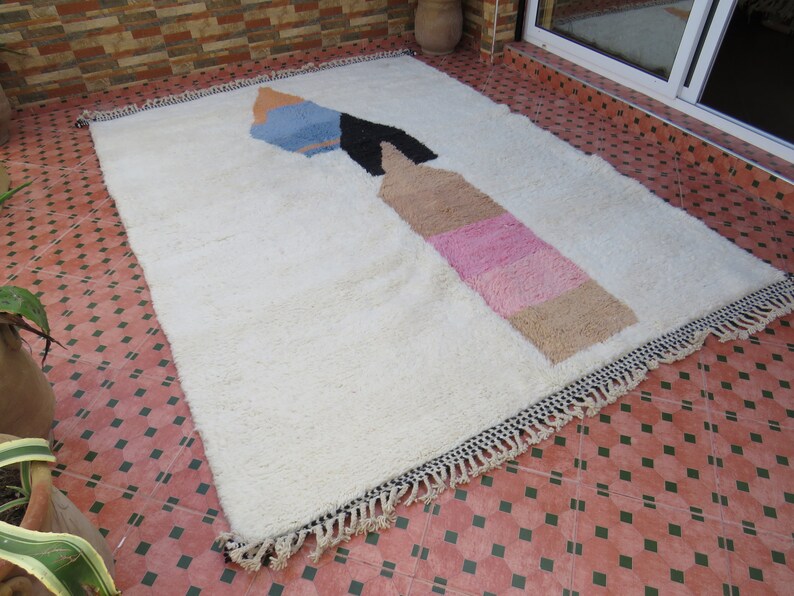 May include: A white shag rug with a colorful abstract design. The design features a large, geometric shape with shades of blue, pink, and brown.