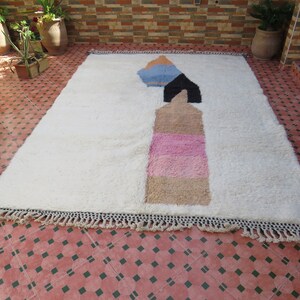 May include: White rug with a geometric design in shades of pink, blue, and brown. The rug has a fringe border.