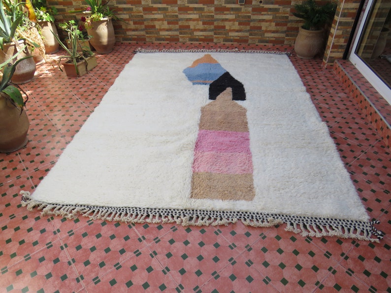 May include: White rug with a geometric design in shades of pink, blue, and brown. The rug has a fringe border.