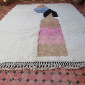 May include: White rug with a geometric design in shades of blue, black, pink, and beige. The rug has a fringe border.