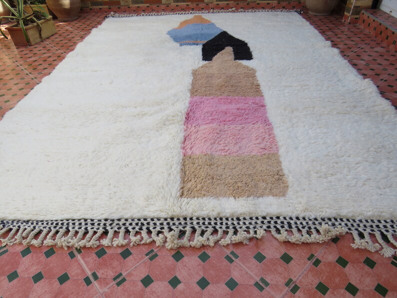 May include: White rug with a geometric design in shades of blue, black, pink, and beige. The rug has a fringe border.