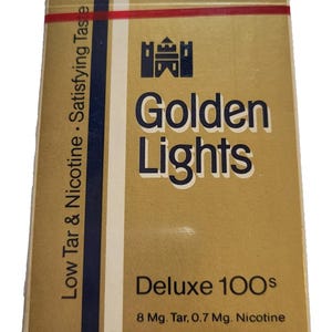 May include: A rectangular box with a gold and white design. The box features the text "Golden Lights" in large, bold letters, with the words "Deluxe 100s" below. The box also includes the text "Low Tar & Nicotine • Satisfying Taste".
