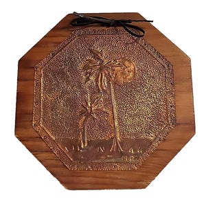 May include: An octagonal wooden wall plaque featuring a central embossed copper panel. The copper depicts two palm trees and a large sun against a textured background. A dark leather cord is attached to the top for hanging purposes.