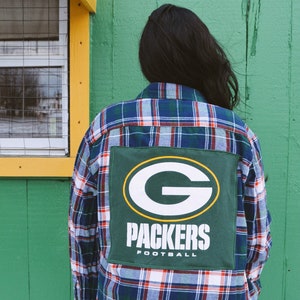 May include: A blue, red, and white plaid flannel shirt with a green patch featuring the Packers logo and the word "PACKERS FOOTBALL" in white. The shirt is worn by a person standing in front of a green wall.