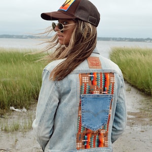 May include: Light blue denim jacket with a colorful patterned back panel and a blue denim pocket. The jacket features a brown patch on the back and is paired with turquoise corduroy pants and a brown trucker hat.