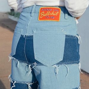 Puede incluir: Vaqueros azules de mezclilla con múltiples capas de tela y bordes deshilachados. Los vaqueros tienen un parche en el bolsillo trasero que dice "Sounds like Bullshit to me".
