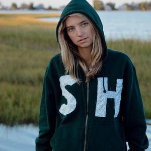 May include: A dark green zip-up hoodie with the letters "SH" in a white, textured font. The hoodie has a hood and a zipper down the front. The background shows a body of water and grassy area.