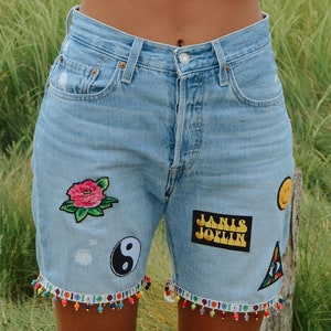 May include: A woman wearing a white tank top, denim shorts with patches and colorful beads, and a white baseball cap with the text "Mother likes you best" in red and blue. She is smiling and holding the hat with both hands.