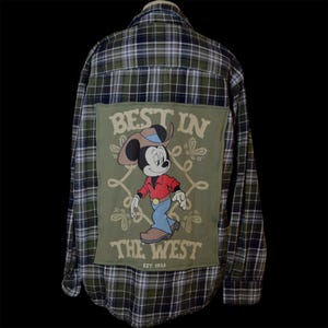 May include: A long-sleeve button-down shirt in a green, navy, and white plaid pattern. The back features a large rectangular olive green patch with a graphic of a cartoon mouse wearing a cowboy hat, red shirt, and blue jeans, with the text BEST IN THE WEST EST. 1925.