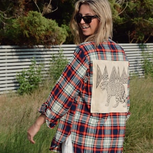 May include: A red, white, and blue plaid flannel shirt with a beige patch on the back featuring a leopard illustration and the text "DA DA DA". The shirt has long sleeves and is worn with white shorts.