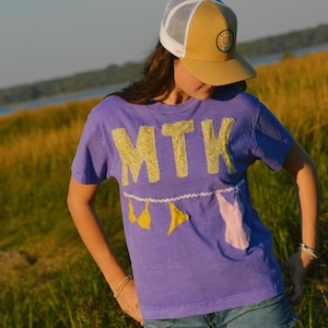 Puede incluir: Una camiseta morada con las letras "MTK" en apliques de tela floral amarilla. La camiseta presenta apliques de bikini amarillos y un aplique rosa. La persona lleva una gorra de camionero amarilla y blanca y pantalones cortos vaqueros.