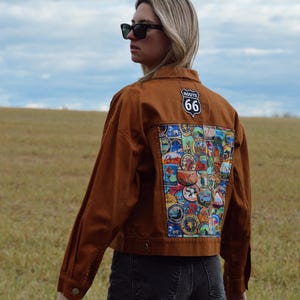 May include: A brown denim jacket with a Route 66 patch and a colorful patch design on the back. The jacket is worn with black jeans. The background is a field and a cloudy sky.