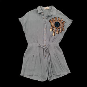 May include: A light gray button-down romper with short sleeves and a drawstring waist. The garment features a large embroidered patch on the upper left chest depicting a stylized eye with gold rays and a dark blue iris, with gold teardrop shapes.