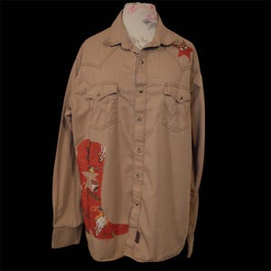 May include: A tan long-sleeve button-down shirt featuring a large red cowboy boot graphic on the lower front, a small red star on the left shoulder, two chest pockets with pointed flaps, and a classic collar with a floral print inner lining.