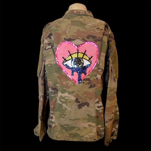 May include: A camouflage print long-sleeve jacket featuring a large, central back patch. The patch is a pink heart shape containing a stylised eye with yellow eyelids, a dark pupil, and blue teardrops, all rendered in shimmering, colourful sequins.