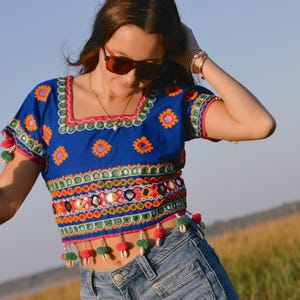 May include: A blue embroidered crop top with a square neckline, adorned with colorful patterns, mirrors, and pom-poms. The top is paired with denim shorts. The overall style is bohemian.