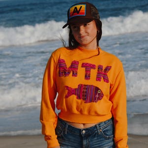 May include: An orange sweatshirt with the letters "MTK" and a fish design, both in a colourful patterned fabric. The person is wearing a brown trucker hat with a colourful "AV" patch and denim shorts. The background is a beach with ocean waves.