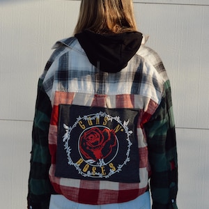 Może przedstawiać: Kurtka z flaneli w stylu patchwork z czarnym kapturem. Kurtka posiada mieszankę wzorów w kratę w kolorach zielonym, czerwonym, białym i czarnym. Na plecach naszywka z logo Guns N' Roses z czerwoną różą i tekstem.