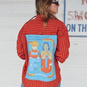 Puede incluir: Una camisa de franela a cuadros rojos y blancos con un parche azul en la espalda. El parche presenta una imagen de dibujos animados de dos niños con las letras "F" y "U" en letras grandes y coloridas. El texto "Today's problems are brought to you by the letters" y "Spelling & Learning" también están en el parche.