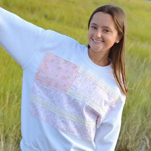 May include: White sweatshirt with a pink and white patchwork design on the front. The patchwork includes floral and paisley patterns, with white lace trim. The model is wearing denim shorts.