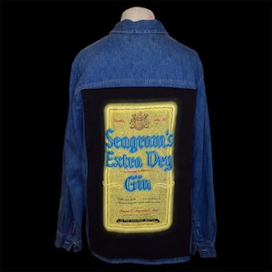 May include: A blue denim long-sleeve shirt featuring a large rectangular black panel on the back. The panel displays a yellow graphic label for Seagram's Extra Dry Gin with blue text, a crest, and the phrase IN THE ANCIENT BOTTLE at the bottom.