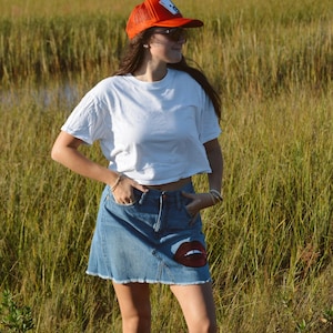 May include: A woman wearing a white t-shirt, denim skirt with a red lips design, and an orange trucker hat. The skirt has a frayed hem. She is wearing black cowboy boots. The background is a field of tall grass.