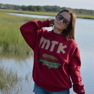 May include: A maroon sweatshirt with the letters "MTK" in a tan, textured font. Below the letters is a colourful, striped design. The person is wearing denim shorts and sunglasses.