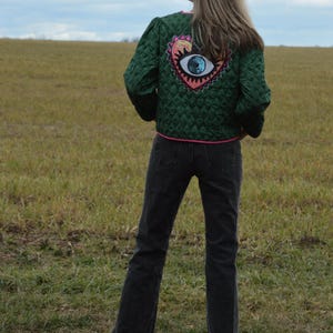 May include: A green quilted jacket with a heart-shaped patch featuring an eye design. The jacket has pink trim and is worn with black jeans. The person is standing in a field of grass.