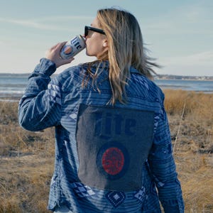 May include: A person wearing a blue denim shirt with a patterned design and a large "Miller Lite" logo on the back. The person is drinking from a can of Miller Lite.