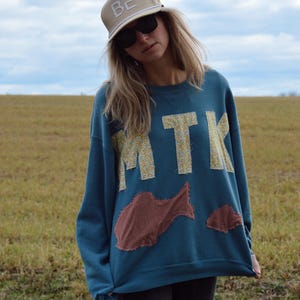 May include: Teal sweatshirt with the letters "MTK" appliquéd in floral fabric. Below the letters are two fish-shaped patches in a red and white checkered pattern. A beige cap with "BE" is worn.