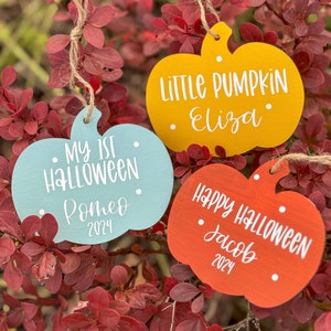 May include: Three wooden pumpkin decorations with painted Halloween messages. The first decoration is light blue with the text "My 1st Halloween Romeo 2024". The second decoration is yellow with the text "Little Pumpkin Eliza". The third decoration is orange with the text "Happy Halloween Jacob 2024".