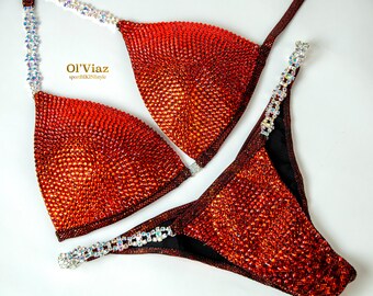 Puede incluir: Un bikini de competición rojo y negro con adornos de cristal. La parte superior del bikini tiene un degradado de cristales rojos, y la parte inferior es negra con un ribete de cristal rojo. El bikini está decorado con tirantes de cristal transparente.