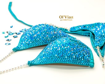 May include: A turquoise bikini top with sparkling blue embellishments. The bikini top is adorned with numerous small, shiny stones, creating a shimmering effect. The straps and edges are accented with silver-colored details. The text "Ol'Viaz sportBIKINIstyle" is visible.