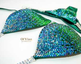 May include: A green and blue bikini top with rhinestones. The top is made of a stretchy fabric and has a halter style strap. The top is decorated with rhinestones in a gradient pattern, starting with blue at the bottom and fading to green at the top. The top is also decorated with a silver chain.
